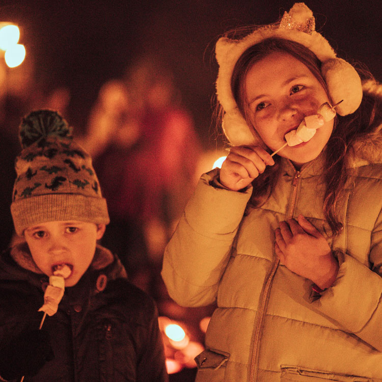 Two children in woolly hats toasting marshmallows by the fire pit
