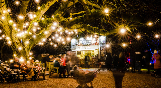Visitors at picnic benches under a tree hung with festoon lights, beside a food stall